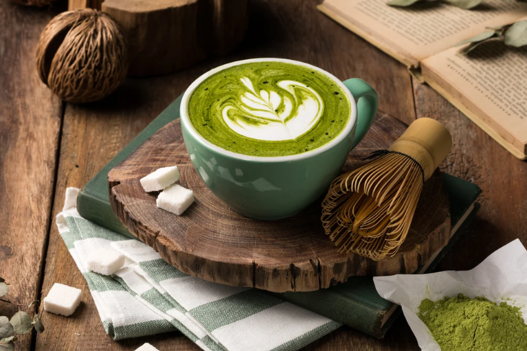 Tazza di matcha latte con decorazione, zucchero e frusta su tavolo di legno.